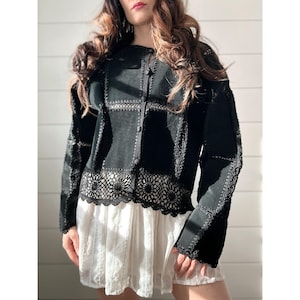 May include: Black suede jacket with crochet detailing. The jacket features a round neckline, button closures, and intricate crochet lace trim along the hemline and sleeves. The jacket is a unique piece of apparel.