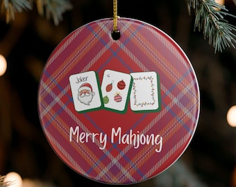Puede incluir: Adorno navideño redondo con un patrón de cuadros rojos y la frase "Merry Mahjong". El adorno presenta tres naipes con diseños festivos, incluyendo un Joker de Papá Noel y adornos navideños.