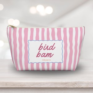 May include: A pink and white striped cosmetic bag with a gold zipper. The bag features the words "bird bam Cheers" in a decorative frame. The bag is made of a soft material and is designed to hold makeup and other beauty products.