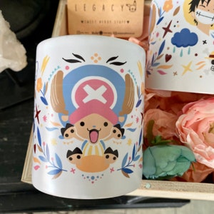 Peut inclure: Une tasse en céramique blanche avec un motif coloré représentant un personnage de dessin animé avec un chapeau rose et un bandana bleu. Le motif comprend des fleurs, des feuilles et des étoiles. La tasse est posée sur une surface en bois.