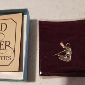 May include: A silver charm depicting a figure on skis holding poles, presented on a dark maroon velvet-like backing inside a clear plastic sleeve, next to a light tan card with black text reading "AND & MMER" and "RSMITHS" inside a blue box.