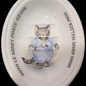 May include: White oval soap dish featuring a cartoon illustration of Tom Kitten in blue clothing. The dish has the text "TOTO LE MINET PORTE-SAVON" and "TOM KITTEN SOAP DISH" around the rim.