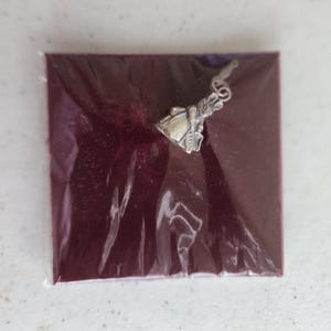 May include: A silver-colored charm in the shape of a bell with a floral design, attached to a small ring. The charm is on a burgundy-colored square, wrapped in clear plastic.