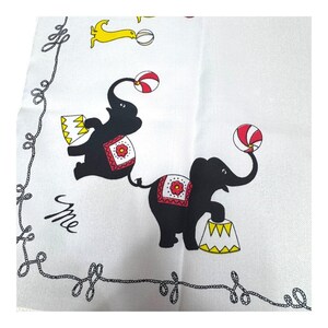 May include: White square textile with a black rope border and circus-themed illustrations. Features black elephants balancing balls and standing on yellow and white platforms. Includes a yellow dog-like figure and the word "me" in cursive.