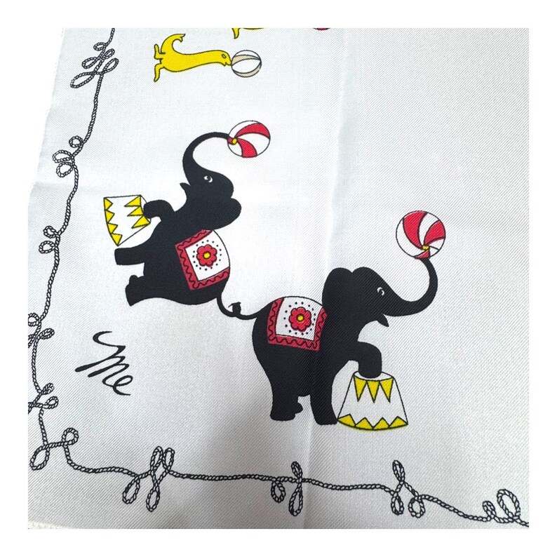 May include: White square textile with a black rope border and circus-themed illustrations. Features black elephants balancing balls and standing on yellow and white platforms. Includes a yellow dog-like figure and the word "me" in cursive.