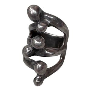 Puede incluir: Un anillo plateado con un dise&ntilde;o abstracto. El anillo presenta una serie de formas redondeadas interconectadas, creando una apariencia &uacute;nica y escultural. El metal tiene una superficie ligeramente texturizada, agregando profundidad al dise&ntilde;o.