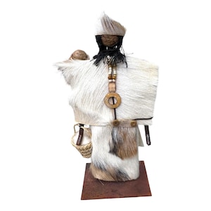 Puede incluir: Muñeca artesanal con una textura similar a la piel blanca y marrón, adornada con un collar de madera y una pequeña cesta tejida. La muñeca se encuentra sobre una base metálica cuadrada de color óxido, mostrando una estética rústica única.