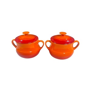 Pode incluir: Duas panelas de cerâmica laranja com tampas e alças. As panelas têm um esquema de cores gradiente, que vai do vermelho profundo nas tampas ao laranja vibrante nos corpos. As alças são curvas e as tampas têm pequenos botões.