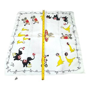 May include: White circus-themed handkerchief with a gray border. The handkerchief features illustrations of circus animals, including elephants, seals, and lions, performing tricks. A yellow measuring tape is placed on the handkerchief, measuring approximately 19 inches.