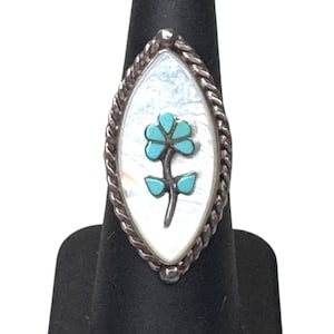 Puede incluir: Anillo de plata con incrustaciones de nácar y diseño floral turquesa. El anillo tiene un borde plateado trenzado y está expuesto en un expositor de anillos negro.