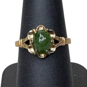Puede incluir: Un anillo dorado con una gran gema verde de forma ovalada. El engaste del anillo tiene un diseño floral alrededor de la gema, con elementos decorativos en la banda. El anillo se exhibe sobre un soporte negro.
