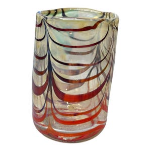 Może przedstawiać: Przezroczysty szklany kubek o unikalnym wzorze. Szkło ma wzór czerwonych, brązowych i czarnych zakrzywionych linii na jasnym tle. Kubek ma lekko nieregularny kształt, co dodaje mu artystycznego uroku.