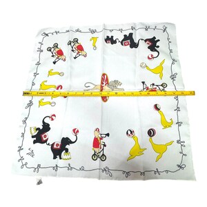 May include: White handkerchief with a circus theme. The handkerchief features illustrations of circus animals, including elephants, seals, and lions, performing tricks. The border has a decorative swirl design. The handkerchief measures approximately 19 inches in length.