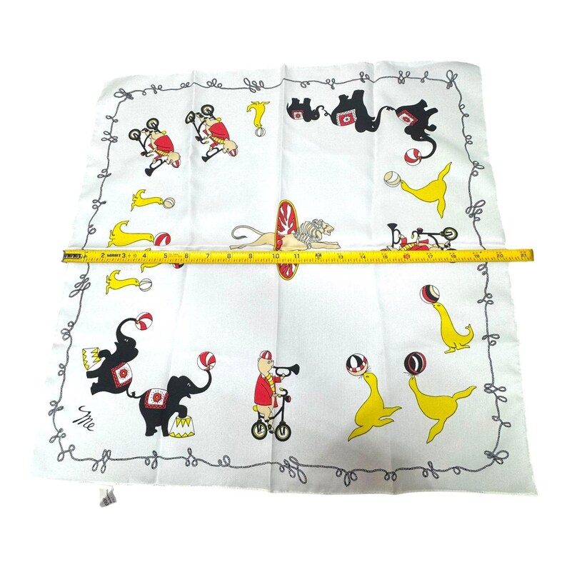 May include: White handkerchief with a circus theme. The handkerchief features illustrations of circus animals, including elephants, seals, and lions, performing tricks. The border has a decorative swirl design. The handkerchief measures approximately 19 inches in length.