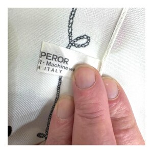 May include: Close-up of a white fabric with a black chain-like pattern. The fabric has a tag that reads "PEROR R-Machine we N ITALY". The fabric is being held by a person's fingers.