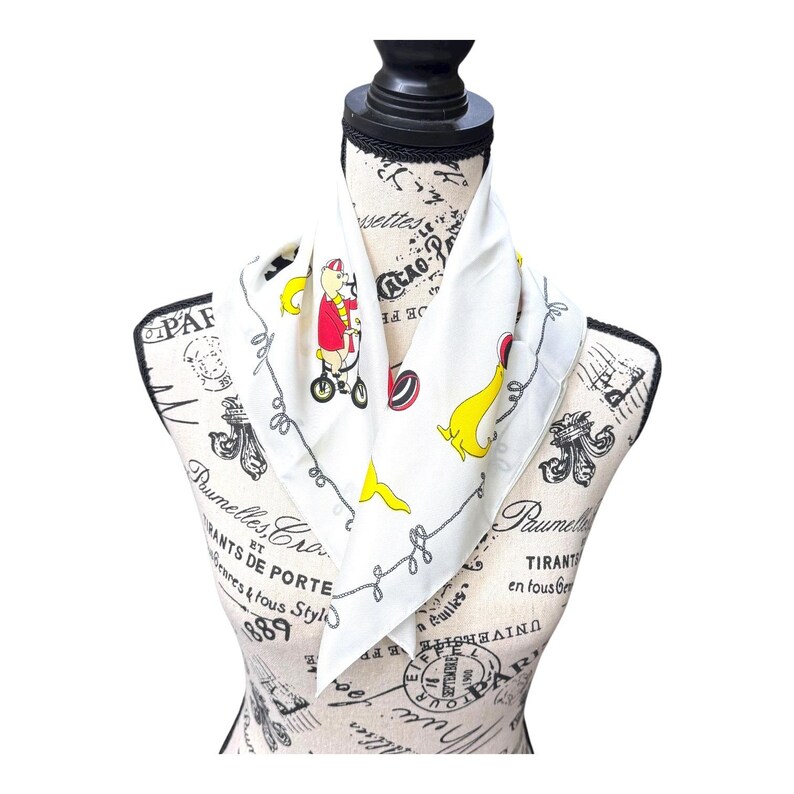 May include: White silk scarf featuring a whimsical cartoon design. The scarf has a repeating pattern of a bear riding a bicycle, yellow bananas, and gray rope-like borders. The scarf is displayed on a mannequin.