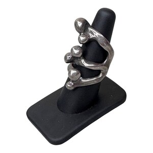 Puede incluir: Un anillo plateado con un dise&ntilde;o abstracto que presenta formas redondeadas y l&iacute;neas curvas. El anillo se exhibe en un soporte negro para anillos sobre un fondo blanco. El dise&ntilde;o del anillo es moderno y escult&oacute;rico.