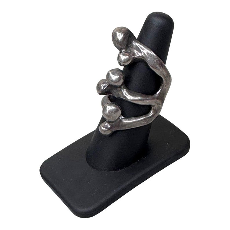 Puede incluir: Un anillo plateado con un dise&ntilde;o abstracto que presenta formas redondeadas y l&iacute;neas curvas. El anillo se exhibe en un soporte negro para anillos sobre un fondo blanco. El dise&ntilde;o del anillo es moderno y escult&oacute;rico.