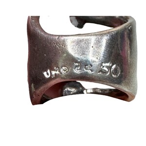 Puede incluir: Primer plano de un anillo plateado con el texto "UNO de 50" grabado en el lateral. El anillo tiene un dise&ntilde;o &uacute;nico y abierto y una superficie ligeramente texturizada. El anillo est&aacute; hecho de un material met&aacute;lico brillante.