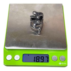 Puede incluir: Un anillo plateado con un dise&ntilde;o abstracto &uacute;nico. El anillo presenta m&uacute;ltiples bandas y acentos esf&eacute;ricos, creando un aspecto moderno y escultural. El anillo se muestra en una b&aacute;scula digital, mostrando un peso de 18,97 gramos.