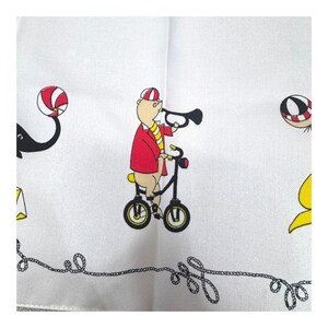 May include: White handkerchief with a whimsical circus theme. A pig in a red jacket and striped hat rides a black bicycle while playing a trumpet. Other circus elements include a black and white ball and a black elephant.