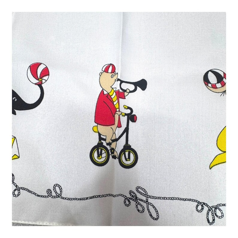 May include: White handkerchief with a whimsical circus theme. A pig in a red jacket and striped hat rides a black bicycle while playing a trumpet. Other circus elements include a black and white ball and a black elephant.