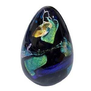 May include: A dark, egg-shaped glass paperweight with iridescent colors. The glass features swirls of green, blue, and gold against a black background. A white, lightning-like design is visible near the top of the paperweight.