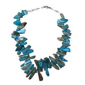 May include: A necklace featuring irregular, turquoise-colored stone chips strung together with a silver-colored chain. The stones vary in shades of blue and green, creating a unique, organic design. The necklace is a statement piece.