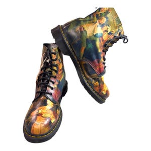 May include: Pair of leather combat boots with a colorful, artistic print. The boots feature black laces, a yellow stitched sole, and a classic lace-up design. The artwork on the boots includes shades of yellow, red, blue, and green.