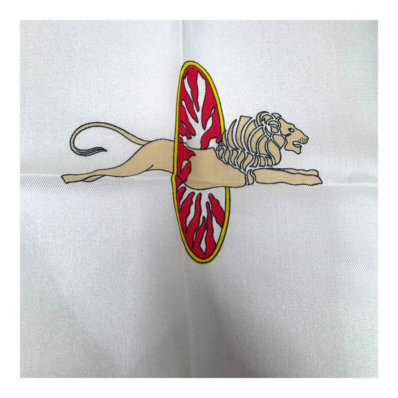May include: White silk scarf featuring a detailed illustration of a lion with a shield. The lion is tan with a cream mane, and the shield is red with a yellow border. The scarf has a subtle texture.