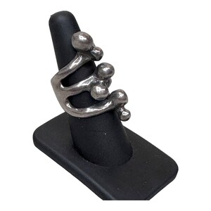 Puede incluir: Anillo plateado con dise&ntilde;o abstracto y m&uacute;ltiples formas esf&eacute;ricas. Se exhibe en un soporte negro, resaltando su forma escultural &uacute;nica y moderna. Una joya original y elegante.