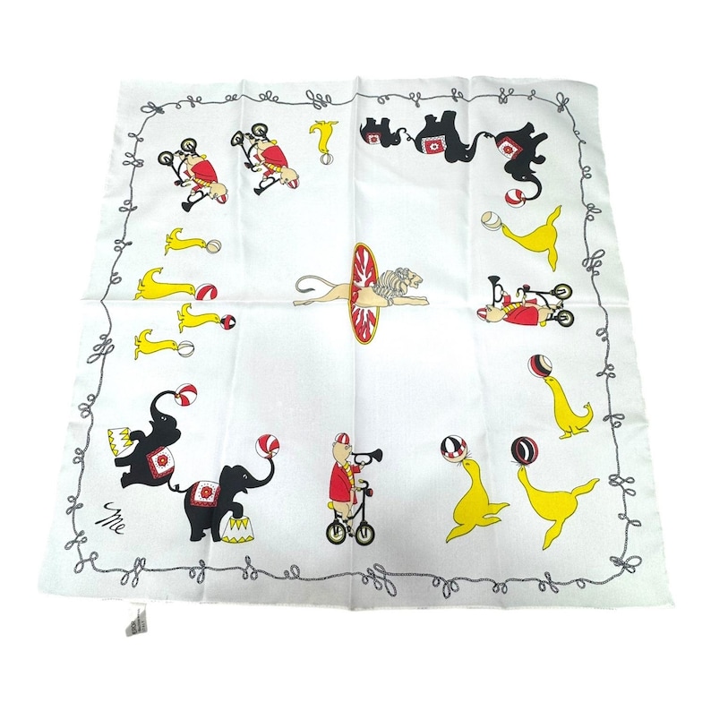 May include: White square handkerchief with a circus theme. The handkerchief features illustrations of circus animals, including elephants, seals, and lions, performing tricks. The border has a decorative gray design.