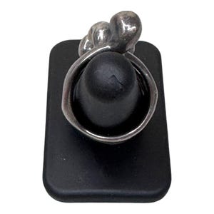 Puede incluir: Un anillo de plata con un dise&ntilde;o &uacute;nico, con tres formas redondeadas en la parte superior. El anillo se exhibe en un soporte rectangular negro, mostrando sus intrincados detalles y artesan&iacute;a. La banda del anillo se curva alrededor del soporte negro.