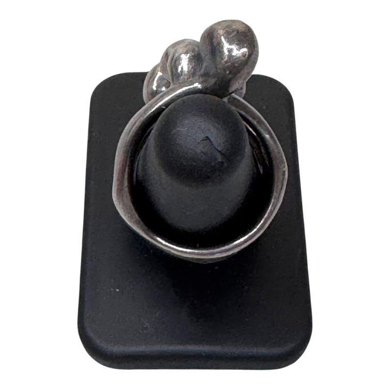 Puede incluir: Un anillo de plata con un dise&ntilde;o &uacute;nico, con tres formas redondeadas en la parte superior. El anillo se exhibe en un soporte rectangular negro, mostrando sus intrincados detalles y artesan&iacute;a. La banda del anillo se curva alrededor del soporte negro.