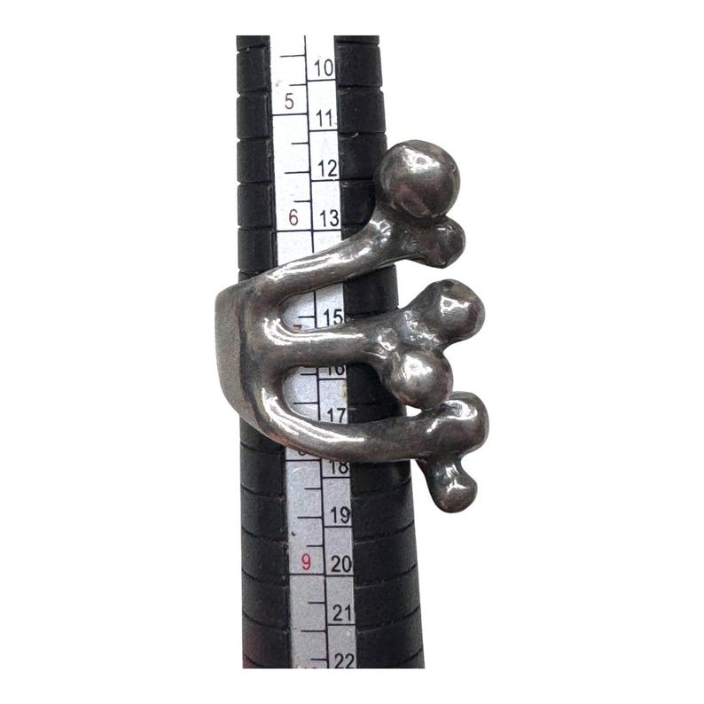 Puede incluir: Un anillo plateado con un dise&ntilde;o abstracto con formas redondeadas y l&iacute;neas fluidas. El anillo se muestra en un medidor de anillos negro, destacando su forma escultural &uacute;nica. El dise&ntilde;o del anillo recuerda formas org&aacute;nicas.