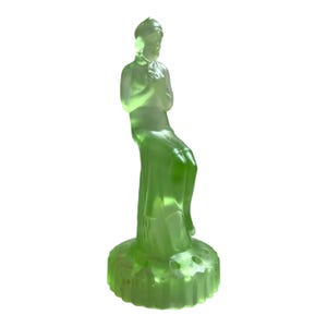 May include: A green glass figurine of a woman sitting on a pedestal. The figurine is made of translucent glass and has a delicate, feminine design.