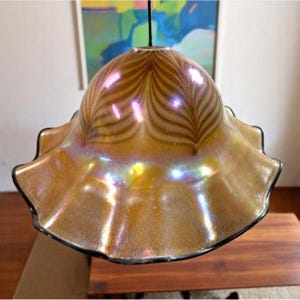 Könnte beinhalten: Eine Vintage-Pendelleuchte aus schillerndem Glas. Die Leuchte hat einen gerüschten, bernsteinfarbenen Schirm mit schwarzem Rand und einem dekorativen Blattmuster. Die Leuchte ist an einem schwarzen Kabel aufgehängt.