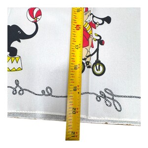 May include: White circus-themed fabric with black and red illustrations. Features a black elephant balancing a ball and a pig riding a bicycle. The fabric measures approximately 21 inches in length.