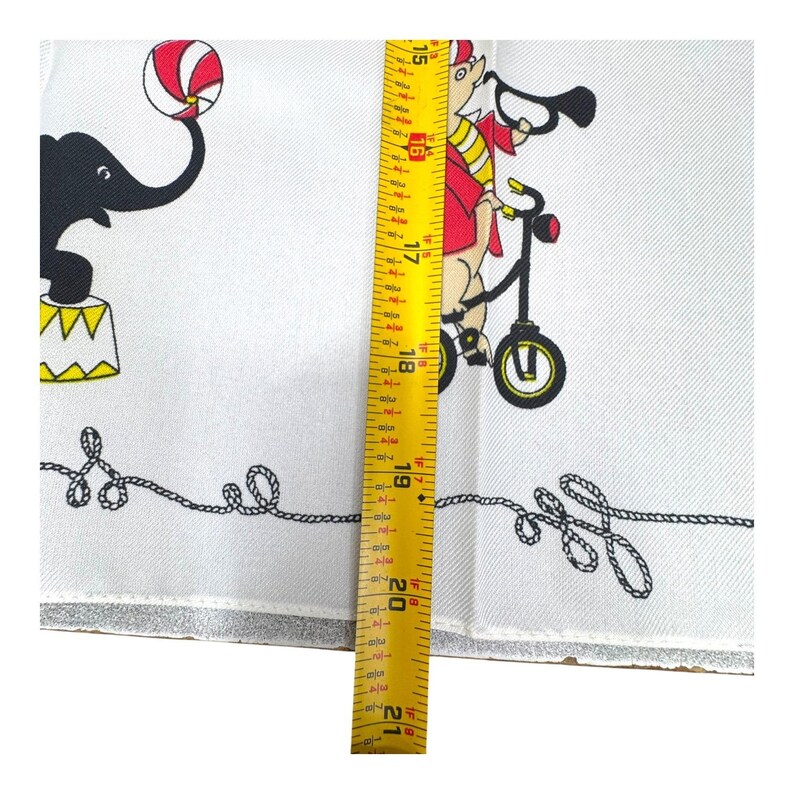 May include: White circus-themed fabric with black and red illustrations. Features a black elephant balancing a ball and a pig riding a bicycle. The fabric measures approximately 21 inches in length.