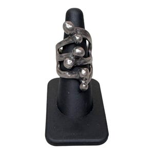 Puede incluir: Un anillo plateado con un dise&ntilde;o &uacute;nico que presenta m&uacute;ltiples elementos redondeados y bandas arremolinadas. El anillo se exhibe en un soporte negro, mostrando sus intrincados detalles y acabado met&aacute;lico. El dise&ntilde;o del anillo es abstracto y moderno.