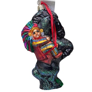 Puede incluir: Adorno navideño de cristal que representa a un niño nativo americano montado a caballo negro. El niño lleva un tocado y ropa coloridos. El caballo es negro con detalles de purpurina y una cinta roja para colgar.