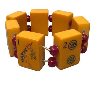 May include: A vintage bracelet featuring rectangular, amber-colored blocks with red beads. Some blocks have black symbols and numbers. The bracelet is strung together with gold-colored wire.