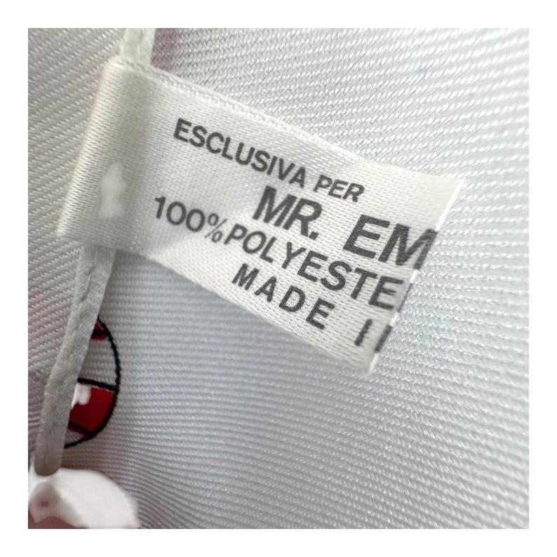 May include: Close-up of a white fabric tag with black text. The tag reads "ESCLUSIVA PER MR. EM 100% POLYESTER MADE IN ITALY". The tag is attached to a white fabric with a subtle texture.