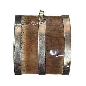 May include: A barrel-shaped pendant with a silver-colored metal frame and bands. The barrel's body has a brown and cream-colored pattern, and a small loop at the top for a chain. The pendant is likely a piece of jewelry.