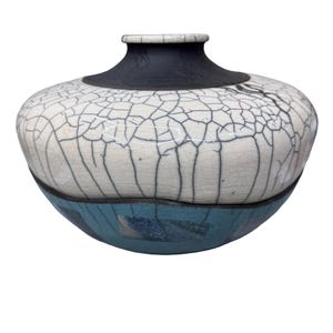 May include: A ceramic vase with a unique crackle glaze design. The vase features a white upper portion with black crackle lines, a teal blue lower section, and a black rim. The vase has a rounded body and a narrow neck.