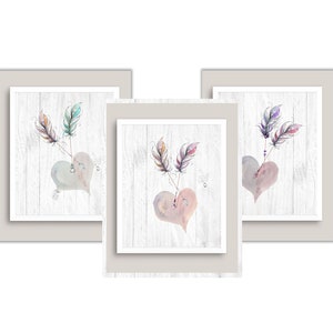 Puede incluir: Un conjunto de tres cuadros de pared con ilustraciones en acuarela de corazones atravesados por flechas con plumas. Cada lámina muestra un fondo de tablones de madera blanca con corazones en tonos pastel y plumas detalladas en rosa, morado y turquesa.