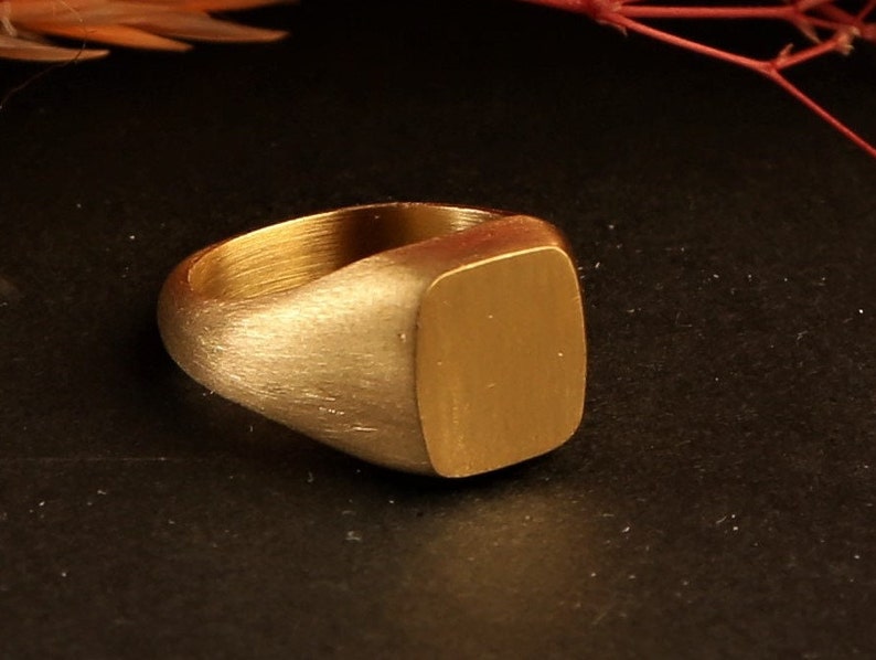 May include: A gold signet ring with a smooth, rectangular top. The ring is made of brushed metal and has a simple, classic design.