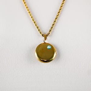 Op de afbeelding: Een goudkleurige ketting met een ronde medaillonhanger. De hanger heeft een kleine blauw-witte boze oog-bedel. De ketting heeft een gedraaid touwontwerp. De ketting wordt getoond tegen een witte achtergrond.