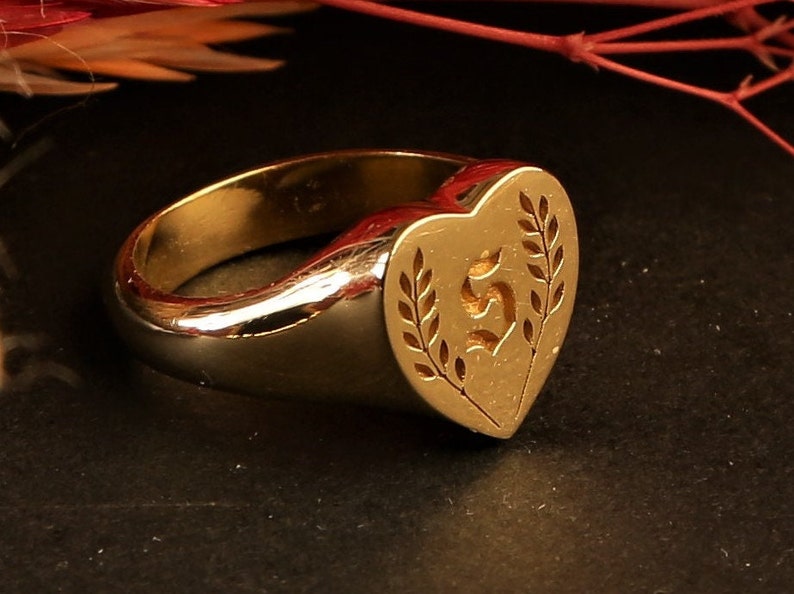 May include: A gold signet ring with a heart-shaped top featuring a stylised letter "S" surrounded by leaves.