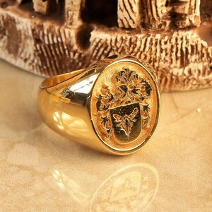 May include: A gold signet ring featuring a detailed crest design. The crest includes a shield with a stylised animal and floral embellishments.